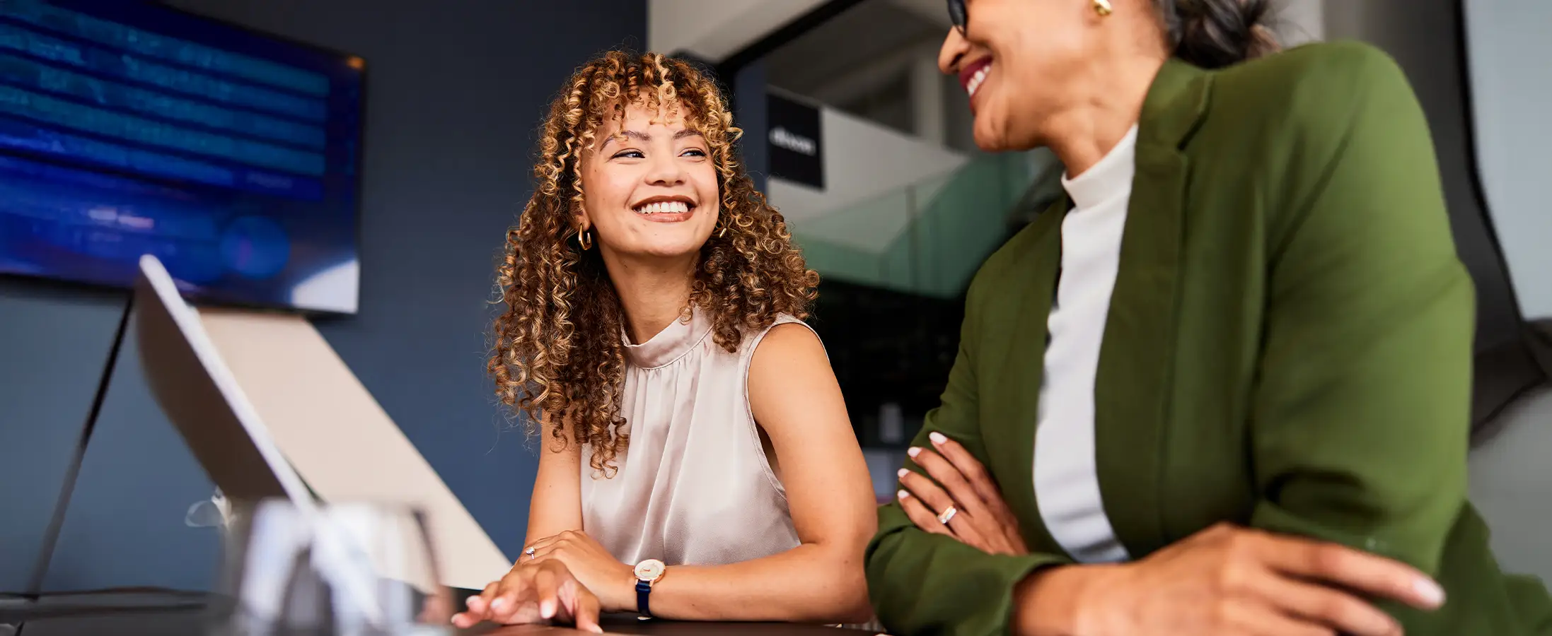 A woman with curly hair a light pink tank top smiles at another woman wearing dark green blazer in an office setting