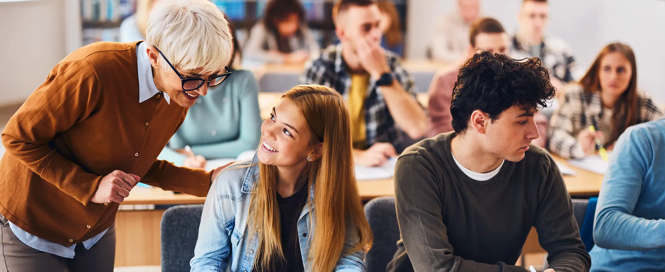 A teacher with glasses and an orange sweater is smiling and talking to a young adult student in a classroom.