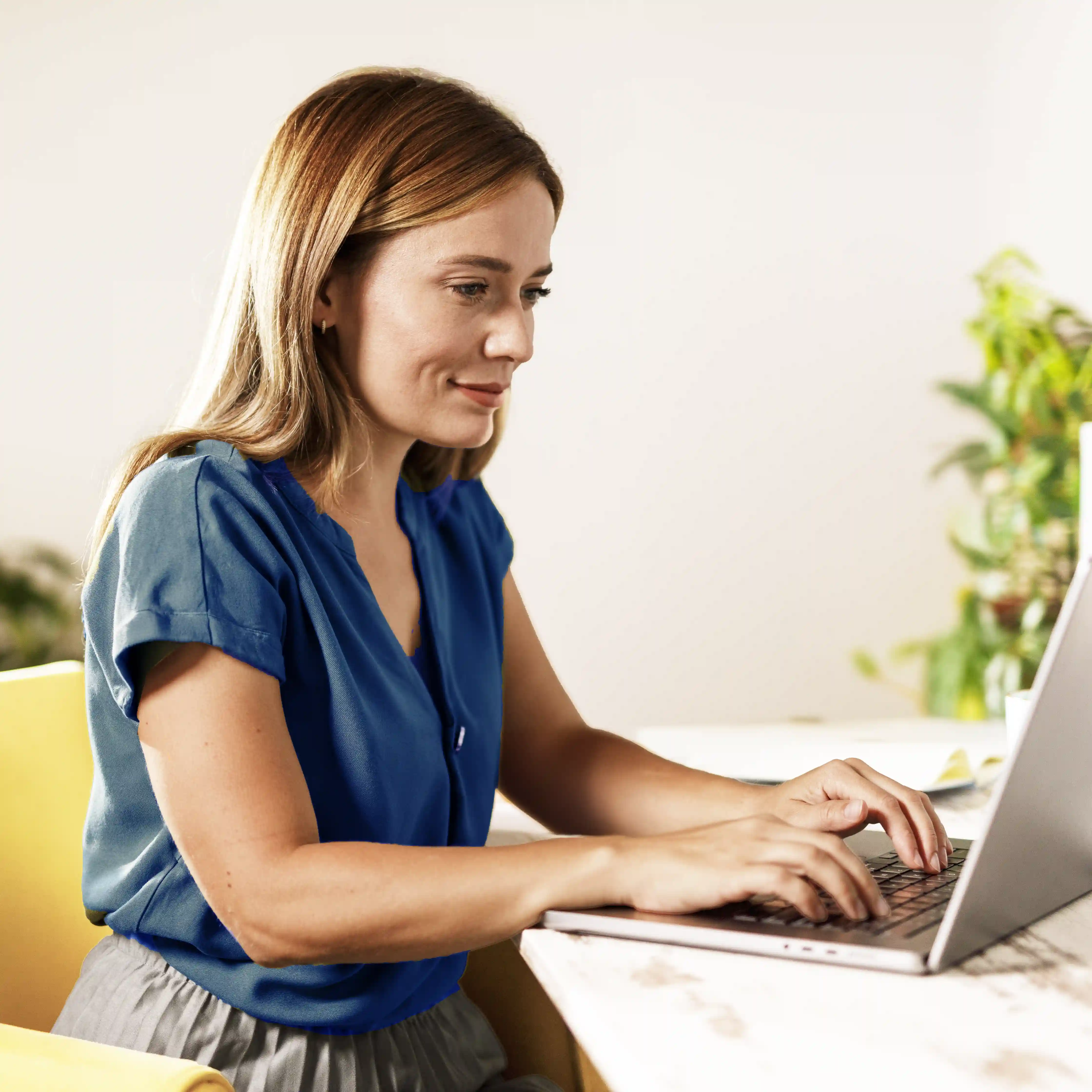 A woman in a blue shirt is typing on her laptop while smiling