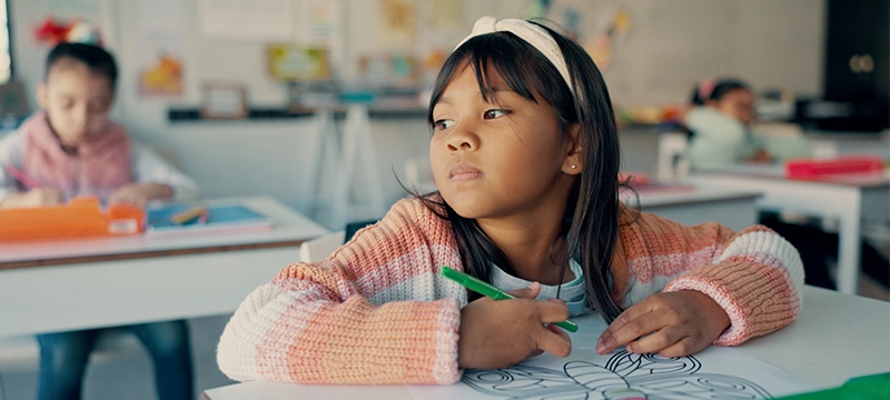 A young girl with black hair looks distracted while sitting in a classroom coloring a picture of a butterfly, symbolizing ADHD and symptoms that parents and teachers should recognize