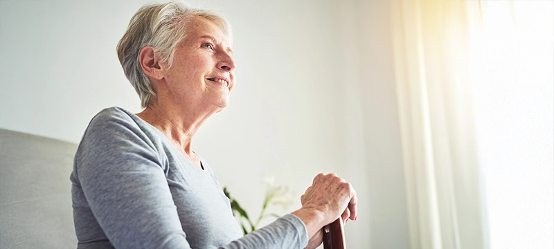 An older woman smiles while sitting, holding her cane, and smiling in a brightly lit room. Representative of Alzheimer's, dementia, and overall brain health.