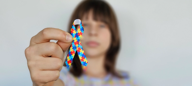 A young child holds up an awareness month bow in a blue, red, and yellow puzzle pattern, symbolizing autism awareness and autism acceptance month