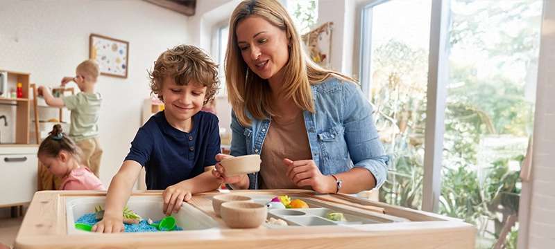 A blonde woman assists a young boy with ASD as he plays in a colored sand pit in a children's playroom or school setting, representing the positive outcomes of using the PDDBI for autism assessment