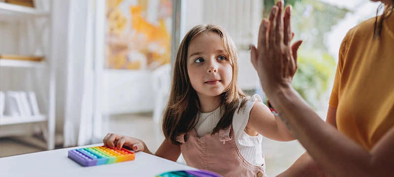 A young girl with autism plays with a rainbow colo9red sensory toy at a table while high fiving a woman to her left, representing social skills and successful autism treatment through using the PDDBI
